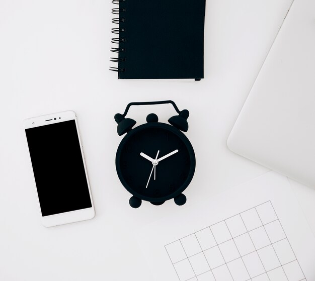 黒の目覚まし時計。スパイラルメモ帳。スマートフォンページと白い机の上のノートパソコン