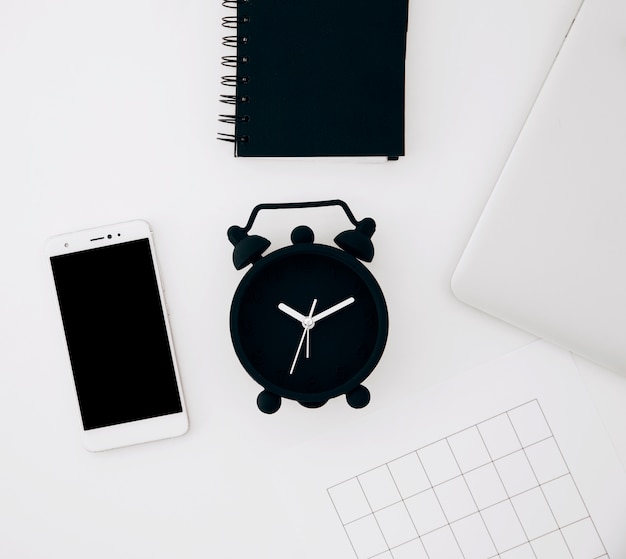 검은 알람 시계; 나선형 메모장; 스마트 폰; 페이지와 화이트 책상에 노트북