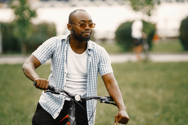 Чернокожий американский мужчина, стоящий на траве со своим велосипедом. Мужчина в белой футболке и синей клетчатой рубашке