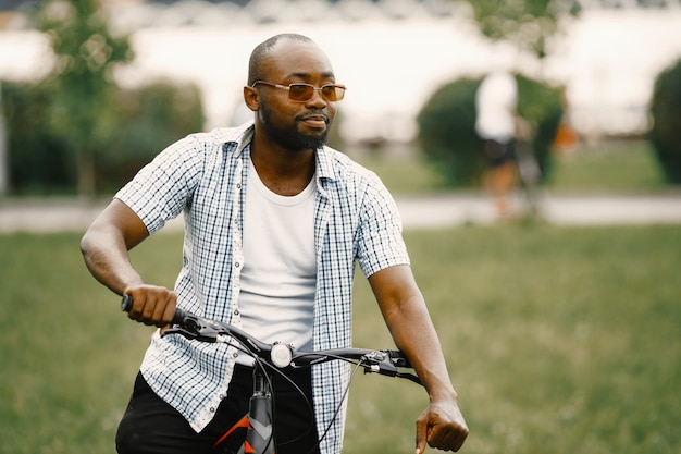 彼の自転車で草の上に立っている黒人のアメリカ人。白いTシャツと青い格子縞のシャツを着ている男