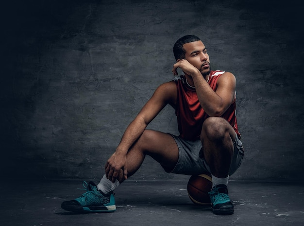 Black basketball player sits on a ball lying on a floor.