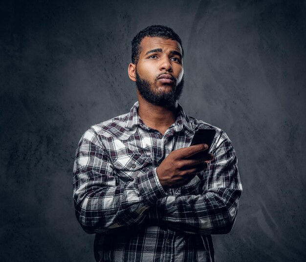 Black bearded male dressed in a fleece shirt using a smartphone and posing over grey background in a studio.