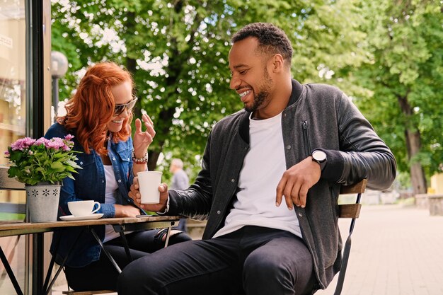 Black bearded male and redhead female drink coffee in a cafe on a street.