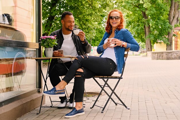 Black bearded male and redhead female drink coffee in a cafe on a street.