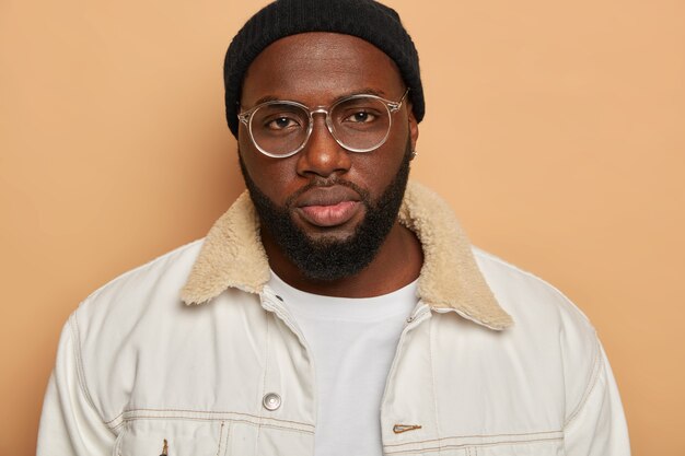  black bearded man has very serious look, wears transparent glasses, black hat and white shirt, looks directly at camera, isolated over beige background. Human facial expressions