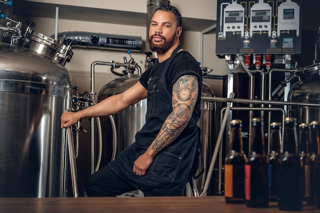 Black bearded tattooed hipster male manufacturer presenting craft beer in the microbrewery.