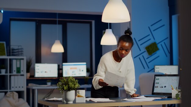 Black bookkeeper working on financial reports checking statistics graphs, looking at laptop, pointing to numbers standing at desk late at night in start-up office doing overtime to respect deadline