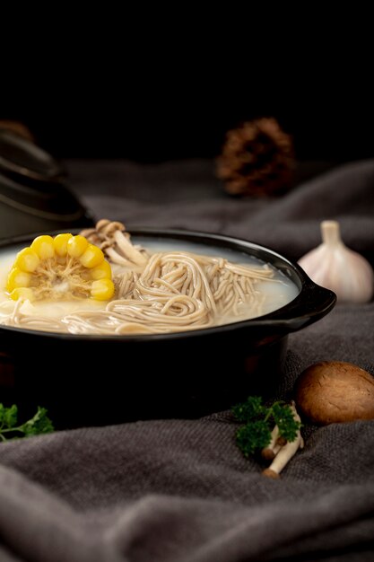 Black bowl filled with noodle soup and corn on a grey cloth with garlic and mushrooms