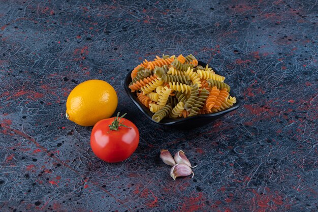 A black bowl full of multi colored macaroni with fresh red tomato and lemon on a dark surface .