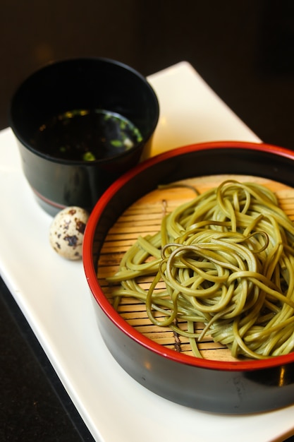 Black bowl of green noodles with quail egg and a sauce served on a white tray