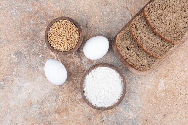 Black bread slices, flour, barley and eggs on marble surface