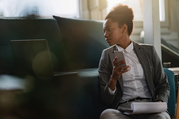 Black businesswoman reading an email on laptop while using mobile phone at home office