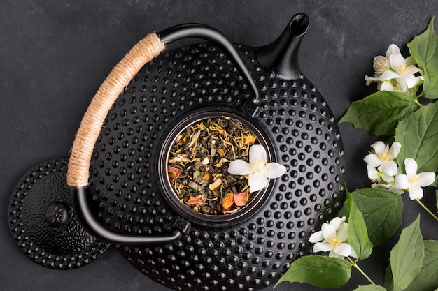 Черный керамический чайник с сухим ингредиентом из травы и веточкой белого цветка на черном фоне