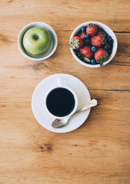 Black coffee cup; green apple; strawberries and blueberries in the bowl on wooden surface