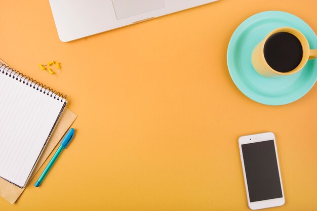 Черный кофе; смартфон; ручка; канцелярская кнопка; ноутбук и блокнот на оранжевой поверхности