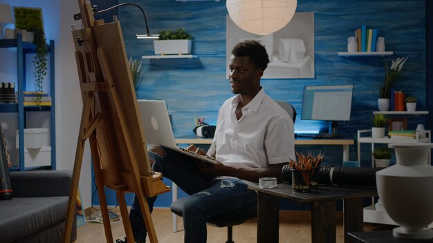 예술 디자인과 걸작을 위한 기술을 사용하는 노트북을 가진 흑인 창의적인 예술가. 성공적인 취미를 위해 연필과 흰색 캔버스로 전문적인 그림을 그리는 아프리카계 미국인