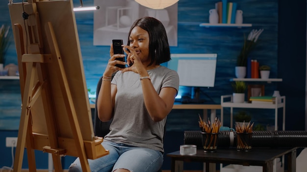 Black creative person holding smartphone for pictures of art drawing in studio space at home. African american young artist using digital technology for modern masterpiece project