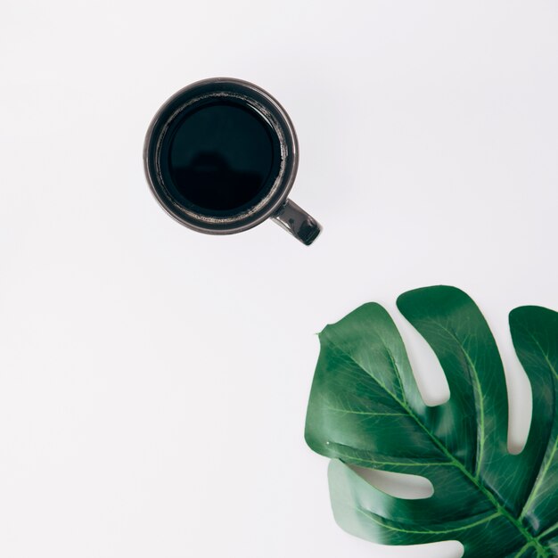 Black cup of coffee and green monstera leaf on white background