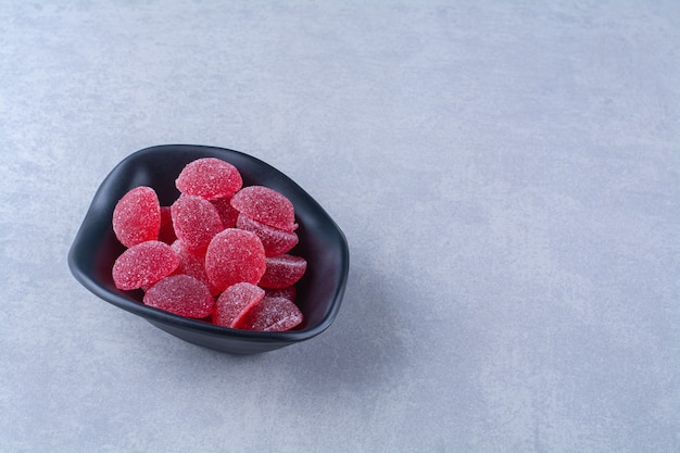 A black deep plate full of red sugary fruit jelly candies on gray table. 