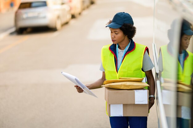 ミニバンの隣に立って、出荷スケジュールリストを読んでいるパッケージを持つ黒人配達の女性