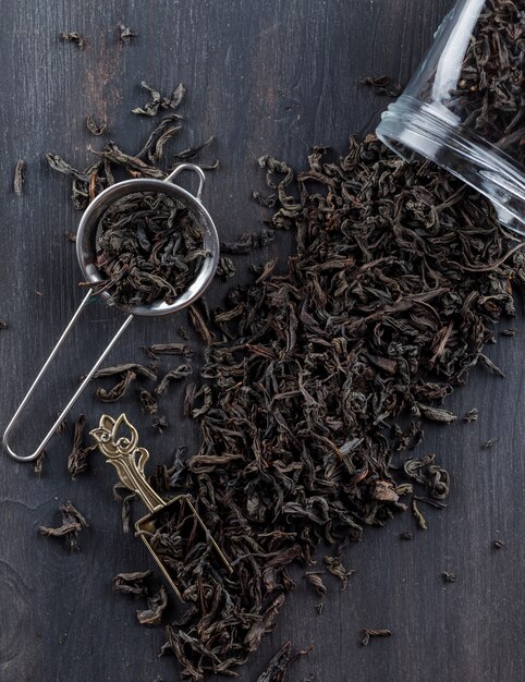 Black dry tea in strainer, jar, scoop on a wooden surface flat lay.