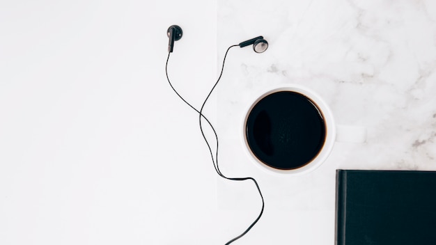 Black earphone; coffee cup and diary on white textured backdrop