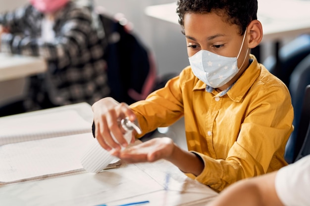 Studente elementare nero che indossa una maschera protettiva e usa un disinfettante per le mani in classe