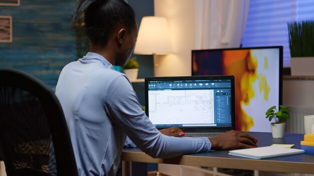 흑인 직원이 밤 늦게 거실 사무실 책상에 앉아 노트북을 보고 있는 회사의 프로젝트를 분석하는 디지털 청사진을 확인합니다. 현대 기술을 사용하는 바쁜 아프리카 프리랜서