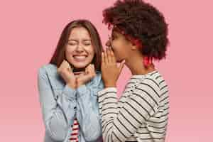 Free photo black ethnic woman whispers secret to her caucasian female with toothy smile, gossip together