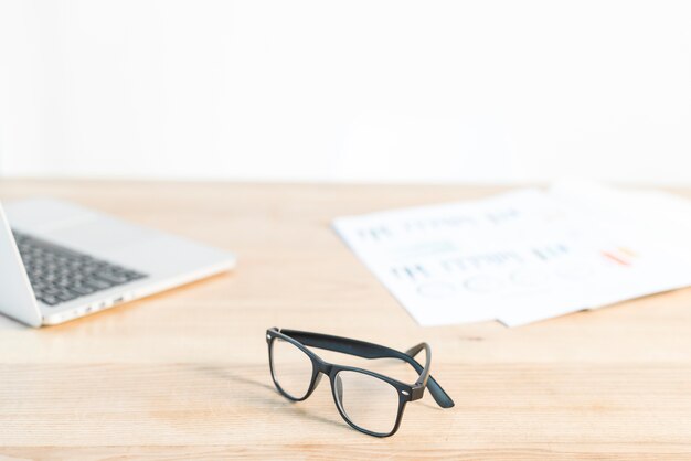 Black eyeglasses in front of laptop and graph on wooden desk