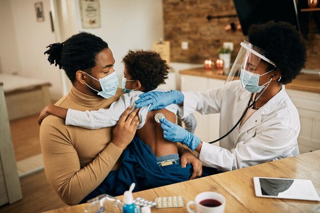 Черная женщина-врач осматривает маленького мальчика стетоскопом во время визита на дом из-за пандемии COVID19