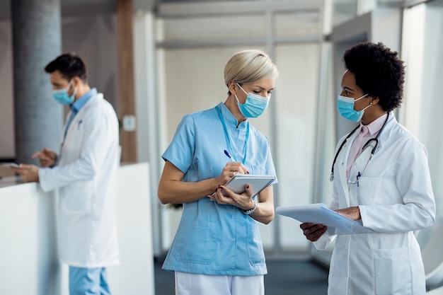 Black female doctor and nurse with face masks communicating at hospital hallway