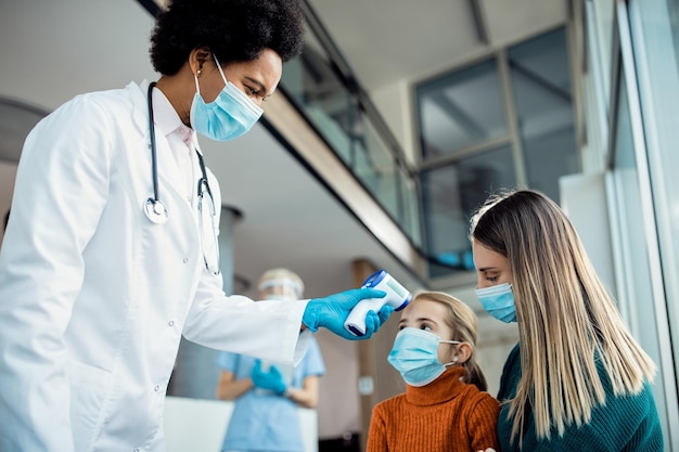 Black female pediatrician measuring temperature of a small girl with infrared thermometer during COVID19 pandemic