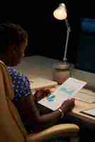 Free photo black female professional sitting at desk in office at night and looking at business chart