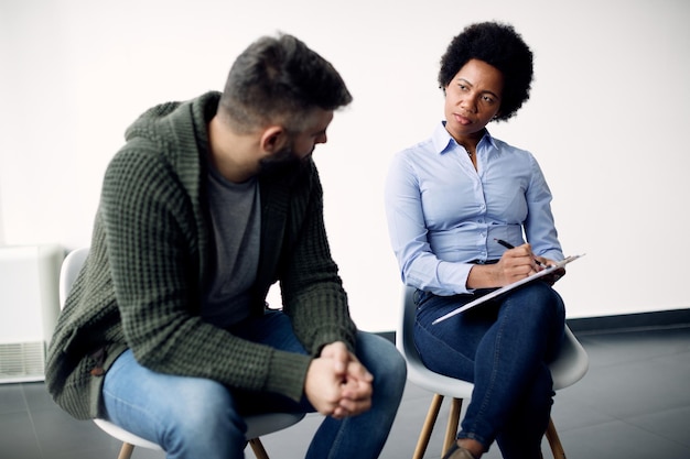 Black female psychotherapist taking notes while talking to a man during counselling