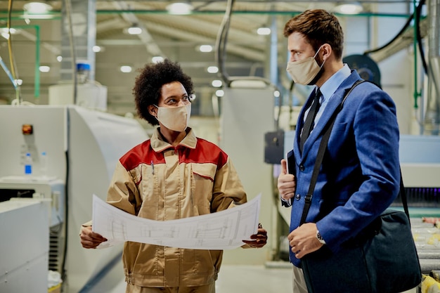 Foto gratuita operaia nera che spiega i piani di progettazione al manager dell'azienda che sta visitando la struttura della fabbrica durante la pandemia di coronavirus