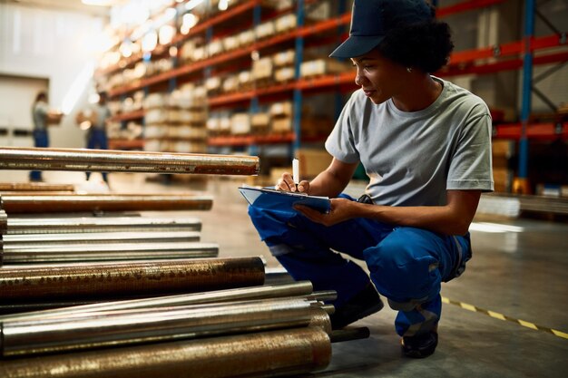 Black female worker writing on clipboard while inspecting steel products at industrial warehouse