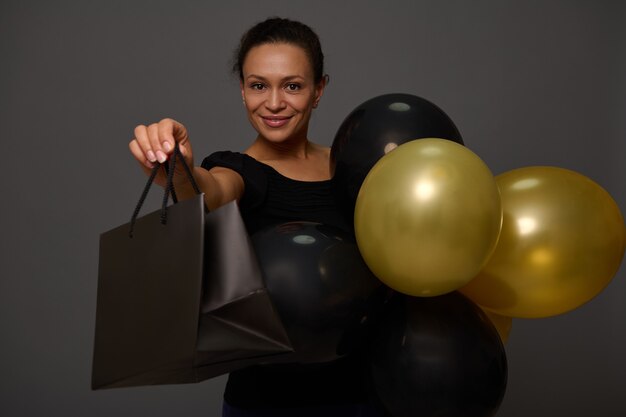 Black friday concept with a cheerful attractive woman with beautiful smile, dressed in black holding black and golden air balloons and shopping bag, looking at camera on background with copy ad space