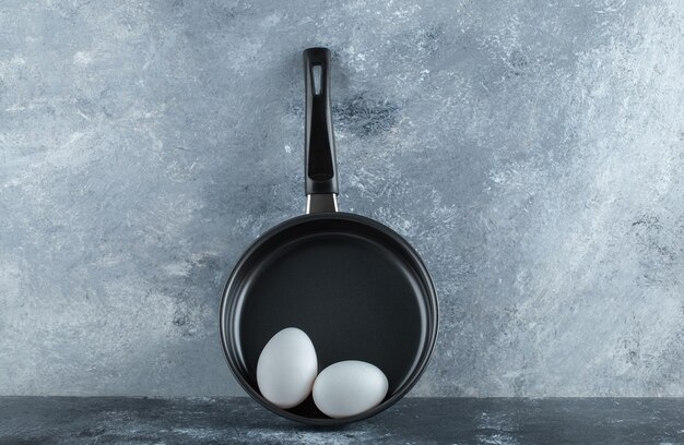 Black frying pan with two organic chicken eggs over grey table.