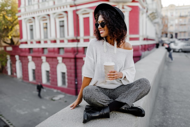 Ragazza nera che si siede sul ponte e che tiene tazza di caffè o tè durante il suo tempo libero. donna freelance. indossa cappello nero e occhiali da sole.