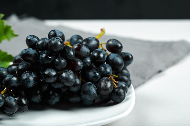 Черный виноград с листьями на белой тарелке с серой скатертью. Фото высокого качества