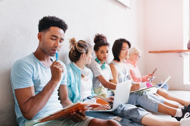 Black guy in blue shirt trying to figure out with difficult test. Stylish female student sitting on the floor with laptop and looking at friends, telling them something.