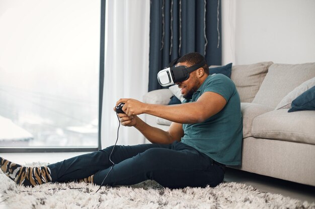 Черный парень в очках виртуальной реальности сидит на ковре в гостиной
