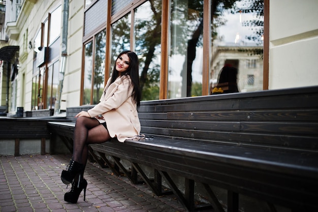 Сексуальная женщина с черными волосами в очках и пальто сидит на скамейке