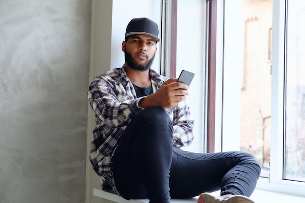 A black hipster male dressed in a fleece shirt and baseball sits near window and using a smartphone.