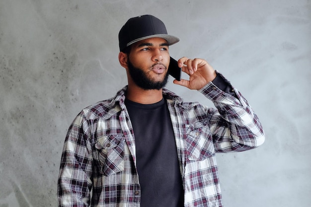 Free photo a black hipster male dressed in a fleece shirt and baseball sits near window and using a smartphone.