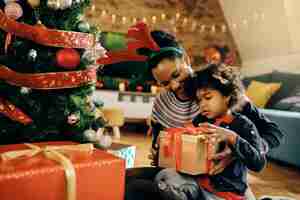 Free photo black little girl and her mother opening gifts by christmas tree at home