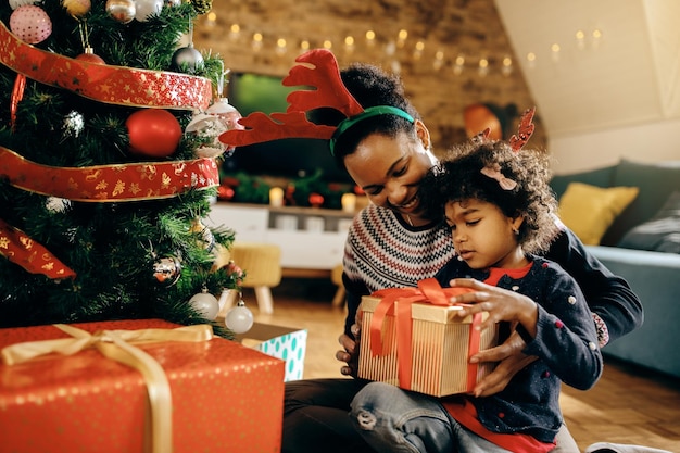 Black little girl and her mother opening gifts by Christmas tree at home