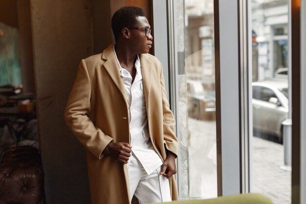 Черный человек в коричневом пальто стоит у окна