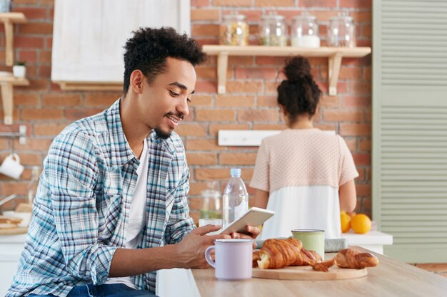 Black man in casual wear checks email or reads world news on electronic device, drinks morning coffee and croissants